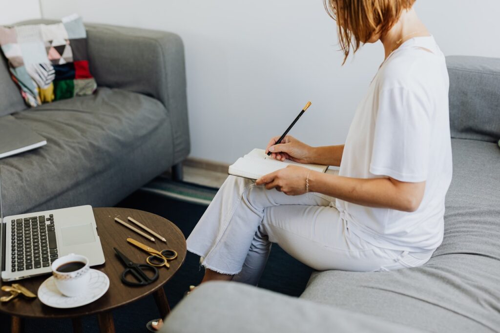En la imagen aparece una mujer sentada en un sofá escribiendo unas notas en un cuaderno junto a su ordenador portátil